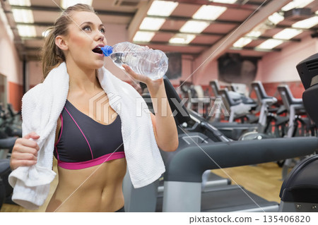 Woman drinking water from clear plastic bottle while pausing treadmill workout in bright gym 135406820