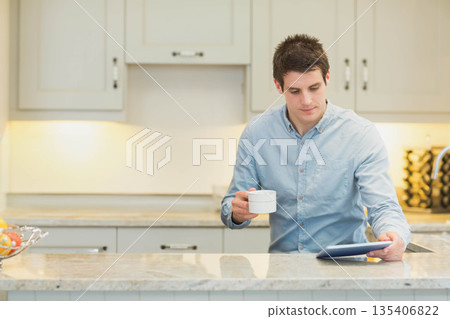 Man standing behind marble countertop in home kitchen holding coffee mug, using tablet, copy space Man standing behind marble countertop in home kitchen holding coffee mug, using tablet, copy space 135406822