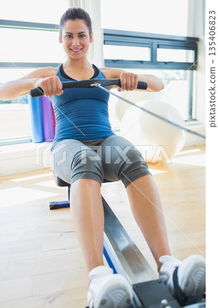 Woman rowing on rowing machine in bright fitness studio with stability ball and yoga mats Woman rowing on rowing machine in bright fitness studio with stability ball and yoga mats 135406823