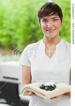 Woman in white uniform holding towel with black stones and white flower in spa, copy space Woman in white uniform holding towel with black stones and white flower in spa, copy space 135406833