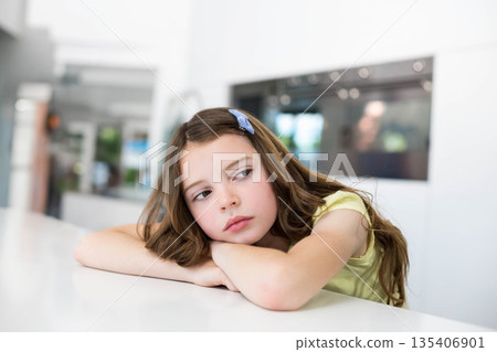 Child girl leaning on white kitchen countertop, wearing blue hair clip, gazing thoughtfully aside 135406901
