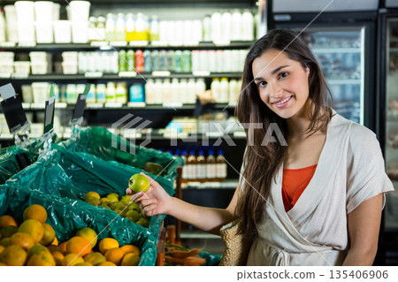 Woman standing in supermarket produce aisle selecting green apple from bin with woven tote bag Woman standing in supermarket produce aisle selecting green apple from bin with woven tote bag 135406906