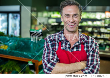 Standing in produce section, male wearing red apron over plaid shirt by wooden bins, copy space Standing in produce section, male wearing red apron over plaid shirt by wooden bins, copy space 135406908