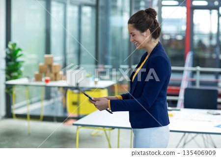 Professional woman in blazer holding tablet, observing office with boxes, laptop, copy space 135406909