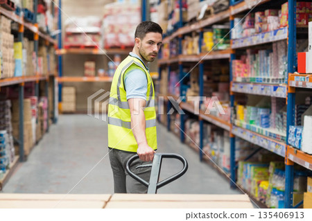 Man wearing safety vest pushing manual pallet jack along warehouse aisle with metal racks and boxes Man wearing safety vest pushing manual pallet jack along warehouse aisle with metal racks and boxes 135406913