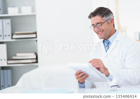Mature doctor in lab coat reviewing clipboard paperwork at office desk with spine model, copy space 135406914