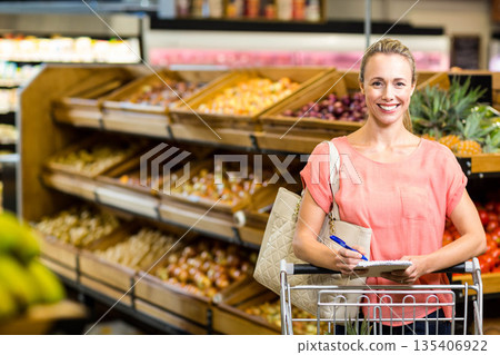 Woman writing notes with pen on notepad by shopping cart in produce aisle, copy space 135406922