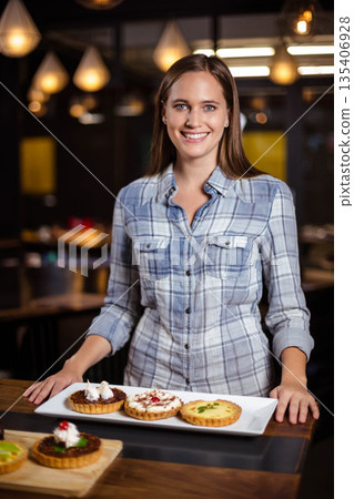 Adult woman in plaid shirt standing behind counter positioning platter with three tarts in bakery Adult woman in plaid shirt standing behind counter positioning platter with three tarts in bakery 135406928
