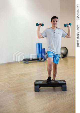 Chinese woman in sportswear stepping onto platform with teal dumbbells in gym, copy space 135406932