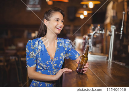 Woman holding brown beer bottle and smiling while sitting at wooden pub bar counter, copy space Woman holding brown beer bottle and smiling while sitting at wooden pub bar counter, copy space 135406936