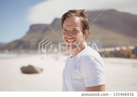 Man standing on sandy beach wearing white t-shirt beside boulder, colorful beach huts, copy space 135406938