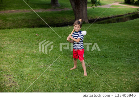 Boy holding baseball bat over shoulder on green lawn at park while baseball flying toward him 135406940