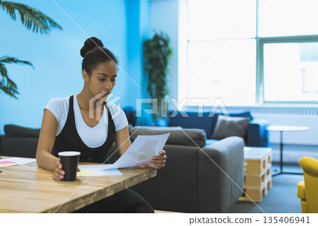 African American woman holding printed documents and sipping coffee at coworking desk, copy space 135406941