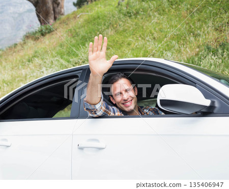 Man wearing plaid shirt waving while leaning out open window of white car at roadside hillside Man wearing plaid shirt waving while leaning out open window of white car at roadside hillside 135406947