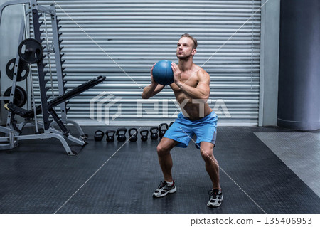 Man squatting holding blue medicine ball at gym interior with kettlebells and barbell rack 135406953