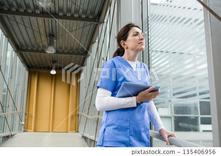 Female nurse in blue scrubs holding tablet near metal railing frosted glass corridor, copy space Female nurse in blue scrubs holding tablet near metal railing frosted glass corridor, copy space 135406956