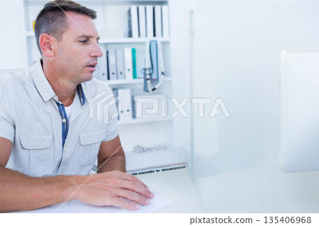 Male office worker sitting at desk in office reviewing documents on computer monitor, copy space 135406968