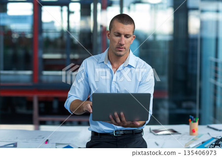 Adult man standing behind desk in modern office typing on laptop among design plans, tablet, pens 135406973