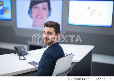 Man in navy blazer turning and smiling at tablet, telephone and screens in control room Man in navy blazer turning and smiling at tablet, telephone and screens in control room 135406997