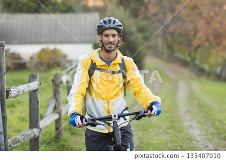 Male cyclist standing beside wooden fence holding mountain bike on rural trail wearing helmet 135407010
