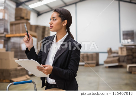 Woman wearing blazer holding clipboard and pen checking boxes on pallets by hand truck in warehouse Woman wearing blazer holding clipboard and pen checking boxes on pallets by hand truck in warehouse 135407013