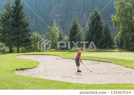 Boy standing in sand bunker holding golf club lining up golf ball at flagstick, copy space Boy standing in sand bunker holding golf club lining up golf ball at flagstick, copy space 135407014