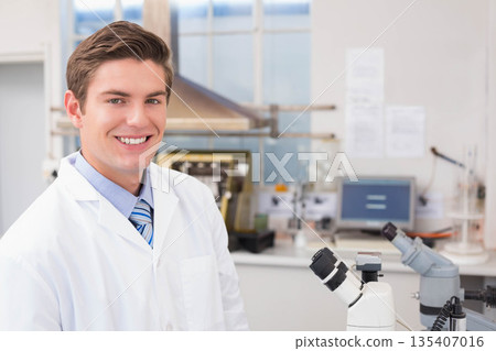 Male researcher standing in lab coat and tie at modern lab bench with microscope, copy space 135407016