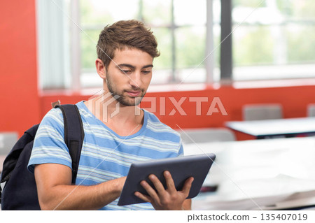 Male college student using tablet in classroom wearing striped tee backpack near desks chairs 135407019