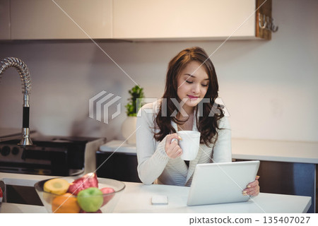 Asian woman sitting at kitchen island holding ceramic mug and looking at tablet near fruit bowl Asian woman sitting at kitchen island holding ceramic mug and looking at tablet near fruit bowl 135407027