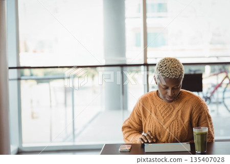 African American woman focusing on tablet while sitting at cafe with green smoothie, copy space 135407028