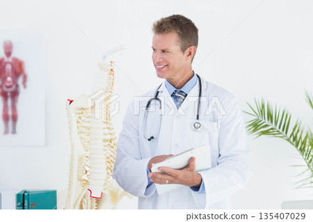 Middle-aged male physician holding clipboard beside spine model and wearing stethoscope in clinic 135407029