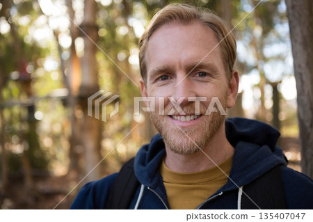 Bearded man standing in sunlit woods wearing navy hoodie and mustard shirt carrying black backpack 135407047