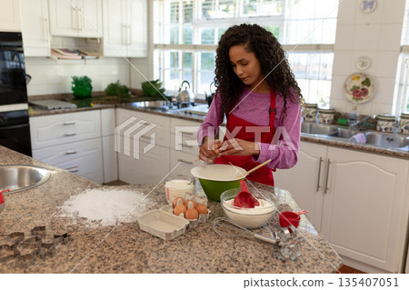 Woman wearing red apron mixing flour and cracking egg into bowls on granite countertop in kitchen 135407051