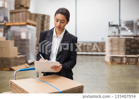 Woman leaning on trolley with cardboard cartons and writing on clipboard near pallets in warehouse 135407054