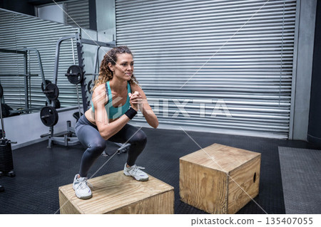 Mid adult woman performing box squat on plyometric box at gym with mirror showing squat rack Mid adult woman performing box squat on plyometric box at gym with mirror showing squat rack 135407055