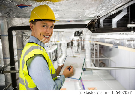 Male engineer wearing helmet, vest checking ducts in hall holding clipboard, pen, copy space 135407057