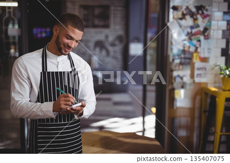 African American male barista wearing apron entering orders using smartphone with stylus in cafe African American male barista wearing apron entering orders using smartphone with stylus in cafe 135407075