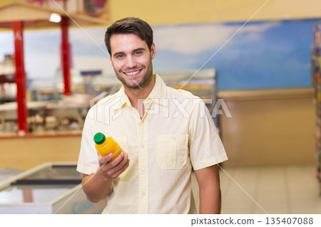 Man holding bottle of orange juice at chest level, standing in store aisle near freezer units 135407088