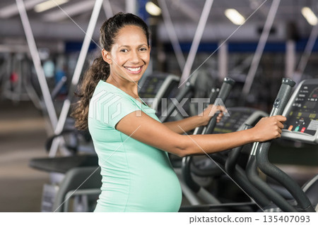 Pregnant woman standing on elliptical machine in gym using console and cardio equipment 135407093