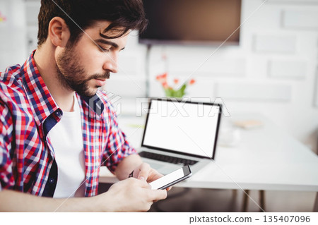 Bearded man scrolling smartphone at desk in home office with laptop, coffee mug, copy space Bearded man scrolling smartphone at desk in home office with laptop, coffee mug, copy space 135407096