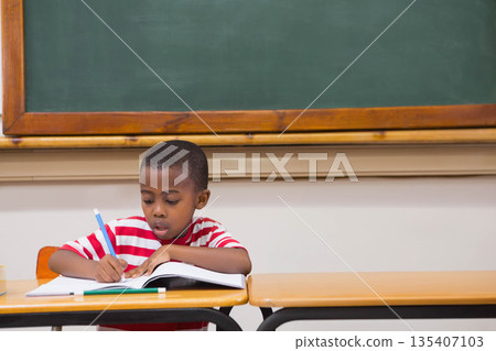 African American boy writing in notebook with pencil at classroom desk with textbook and chalkboard African American boy writing in notebook with pencil at classroom desk with textbook and chalkboard 135407103