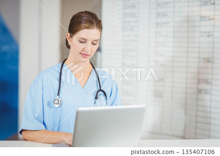 female nurse typing on silver laptop behind reception counter, reviewing wall chart data 135407106