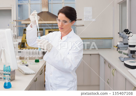 Female scientist at lab bench wearing lab coat pouring blue solution with pipette into test tube Female scientist at lab bench wearing lab coat pouring blue solution with pipette into test tube 135407108