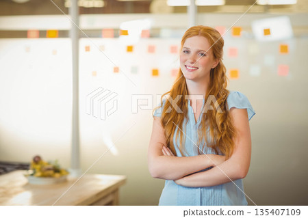 Businesswoman standing with arms crossed in office by glass partition with sticky notes and bananas 135407109