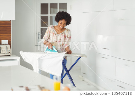African American adult woman pressing white shirt on ironing board in kitchen with turquoise iron 135407118