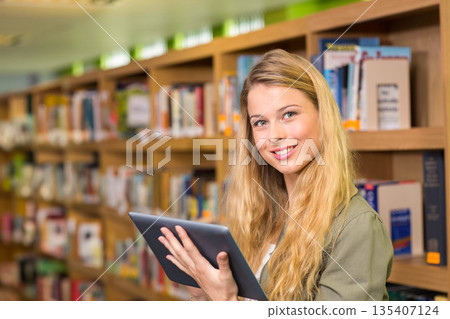 Teenage girl standing in library aisle holding tablet and glancing toward camera, copy space Teenage girl standing in library aisle holding tablet and glancing toward camera, copy space 135407124