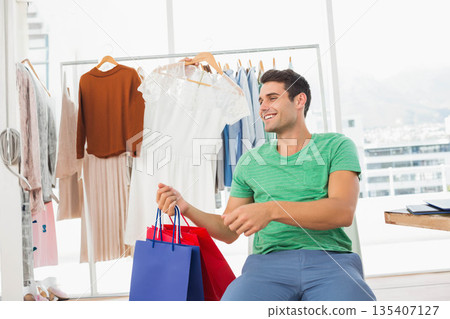 Adult male shopper sitting on bench holding red and blue paper bags in showroom, copy space Adult male shopper sitting on bench holding red and blue paper bags in showroom, copy space 135407127