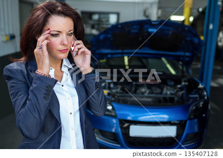 Woman talking on smartphone beside blue car with open hood at workshop, vehicle lift, copy space 135407128