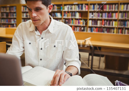 Male student studying in library reading room at wooden table with laptop, open books, copy space Male student studying in library reading room at wooden table with laptop, open books, copy space 135407131