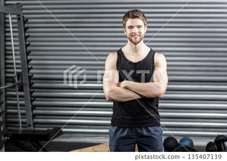 Muscular man folding arms and smiling toward camera inside gym area with squat rack and kettlebells 135407139